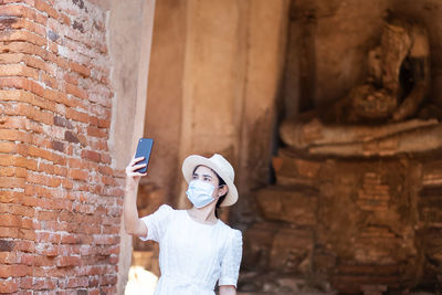 Mid adult woman wearing mask doing selfie while standing against ancient structure