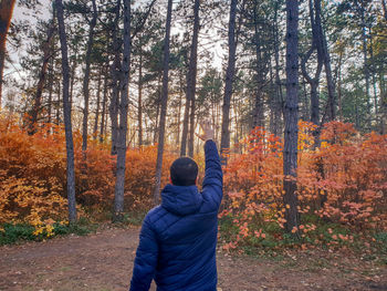 Rear view of young man with hand raised standing amidst trees in forest during autumn