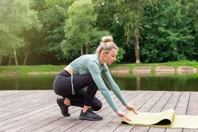 A woman in sportswear, in summer, by pond, unwinds a green gym mat, prepares for sports.