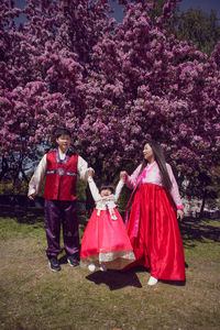 Korean family in national costumes in nature stands next to a cherry blossoming tree.