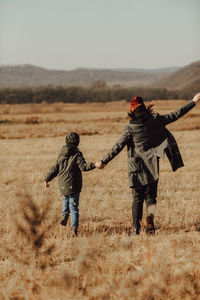 Child in warm green knitted hat and his mother holding hands and running through the autumn field 