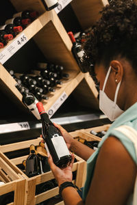 Side view of crop african american female customer in protective mask for coronavirus prevention examining bottle of wine while choosing drinks in supermarket