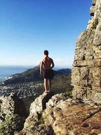 Full length rear view of shirtless climber standing on cliff against clear blue sky
