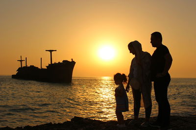 Silhouette family on shore against sky during sunset