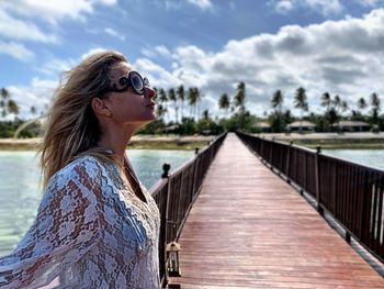 Woman wearing sunglasses while standing on footbridge over river against sky