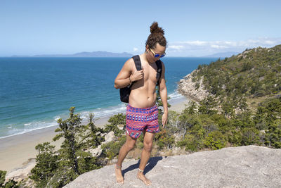 Full length of shirtless man standing on rock by sea against sky