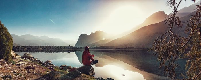 Man looking at view while sitting on rock by lake against mountains and sky during sunset