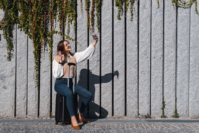 Young cheerful redhead woman with glasses sitting on black suitcase while taking selfie