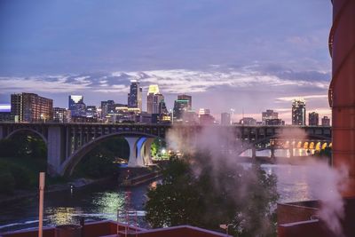 Illuminated bridge over river by buildings against sky during sunset