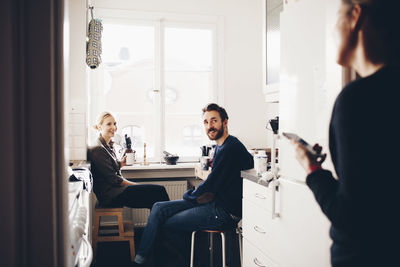 Happy couple sitting in kitchen while looking at female friend