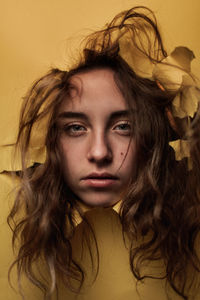 Pensive happy youthful female teen with uncombed hairstyle watching out from ripped hole in yellow paper in studio