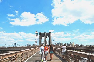 People walking at brooklyn bridge against cloudy blue sky on sunny day