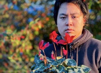 Portrait of young asian man holding red flowering cyclamen plant against trees.