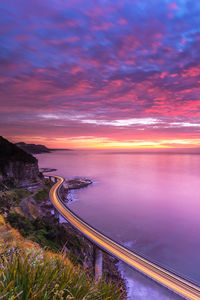 Light trails on road by sea against sky at sunset