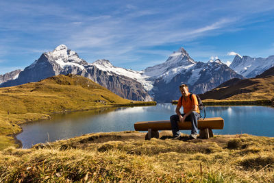 Man sitting on bench against lake with mountains and sky