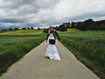 Woman standing on footpath amidst grassy field against cloudy sky