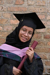 Portrait of young woman standing against wall with convocation suit during her graduation