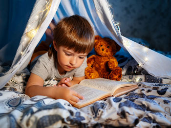 Little boy reads book with teddy bear. toddler plays in tent made of linen sheet on bed. coziness.