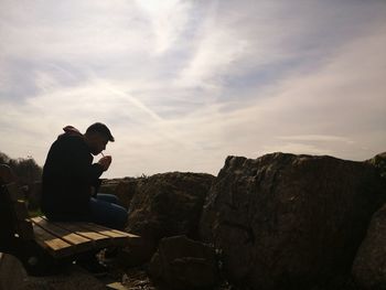 Man sitting on bench smoking cigarette against sky