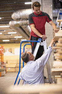 Male customer showing plank to salesman standing on ladder at hardware store