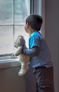 A young boy with his stuffed bear, looks out the window, watching for grandma