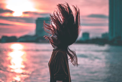 Side view of young woman standing by sea against sky during sunset