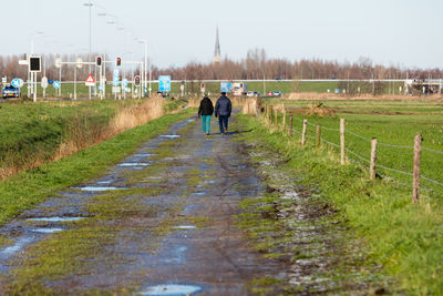 Rear view of people walking on dirt road amidst field