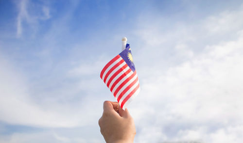 Cropped hand of person holding malaysian flag against cloudy sky