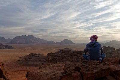 Rear view of man sitting at desert against sky during sunset