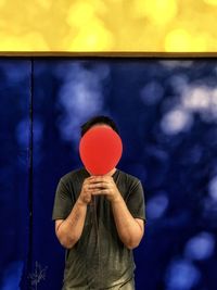 Midsection of man holding red balloon against blue and yellow wall.
