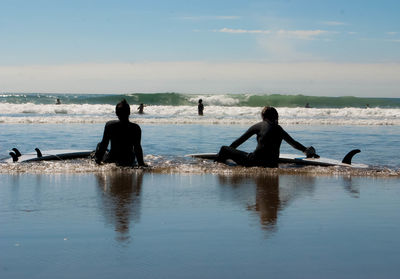 Rear view of friends sitting on shore at beach against sky