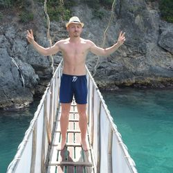 Portrait of shirtless man wearing hat standing on footbridge over sea