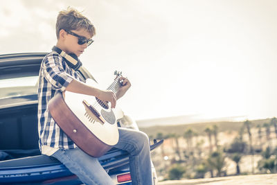 Young boy playing acoustic guitar sitting on the car trunk outdoors. musician singing on the road