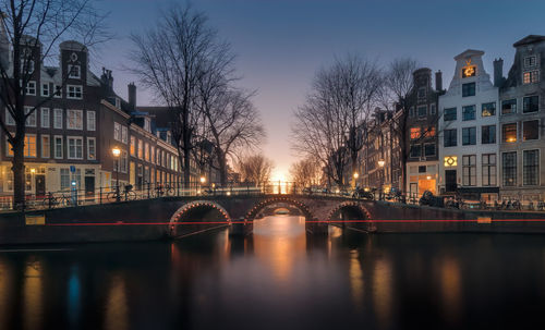 Bridge over river by illuminated buildings against sky at night