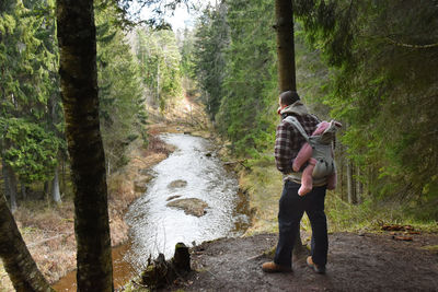 Dad carrying his 7 month old baby girl on his back in ergo bag in the woods, river in background