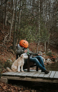 Man wearing scary pumpkin head for halloween sitting on dock with dog.