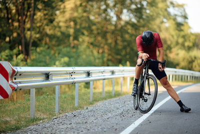 Tired road cyclist in helmet and sport clothing resting on bike among green nature. 