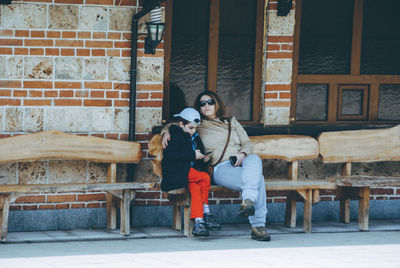 Full length of woman sitting with boy on bench against building