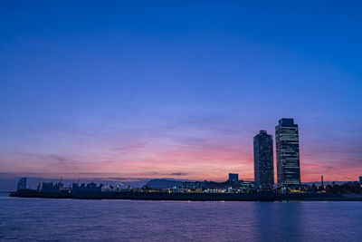 Sea by modern buildings against sky during sunset