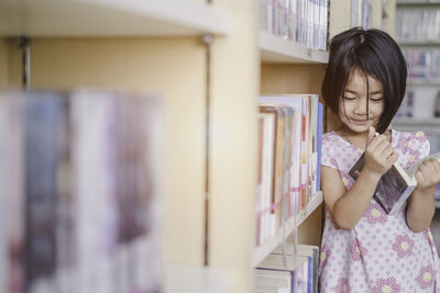 Girl looking away while holding book by shelves in library