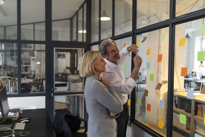 Happy colleagues looking at sticky notes at glass pane in office