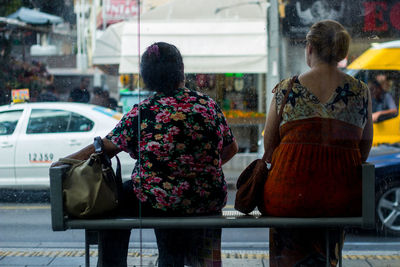 Rear view of women sitting on bench in city seen through glass