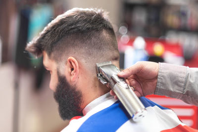 Cropped hand of barber cutting male customer hair with clipper in salon