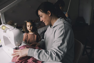 Daughter looking at mother sewing clothes at home