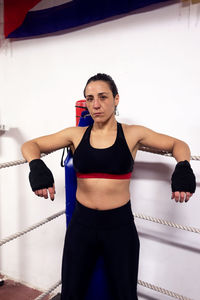 Real female boxer in gloves leaning on rope at ring before training