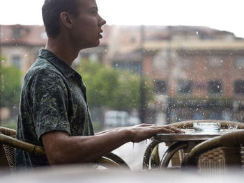 Side view of young man sitting at sidewalk cafe during rain