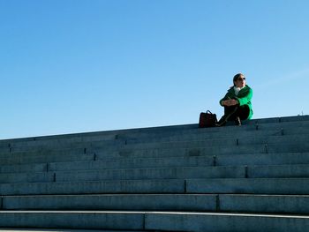 Low angle view of woman sitting on staircase against clear blue sky