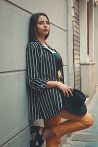 Model standing against  wall wearing a dark grey jacket black hat yellow pants looking at camera.