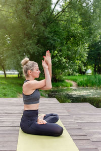 A beautiful woman sitting on a wooden platform by a pond on green mat in summer, does yoga