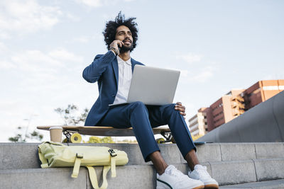 Spain, barcelona, young businessman sitting outdoors in the city using cell phone and laptop